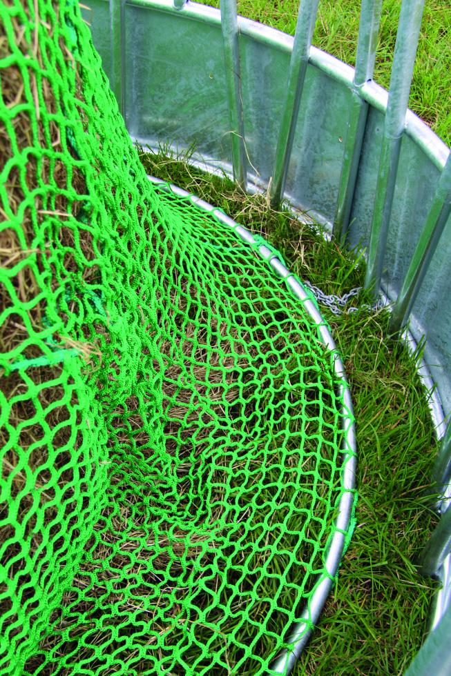 Eine Detailansicht einer runden Rundbogengitterraufe auf Gras, grünes Futtersparnetz überzieht teilweise sichtbarer Futterhaufen und ist an Spannrahmen befestigt, welcher auf dem Boden liegt, Ansicht von oben, zeigt die Hälfte des Innern der Raufe 